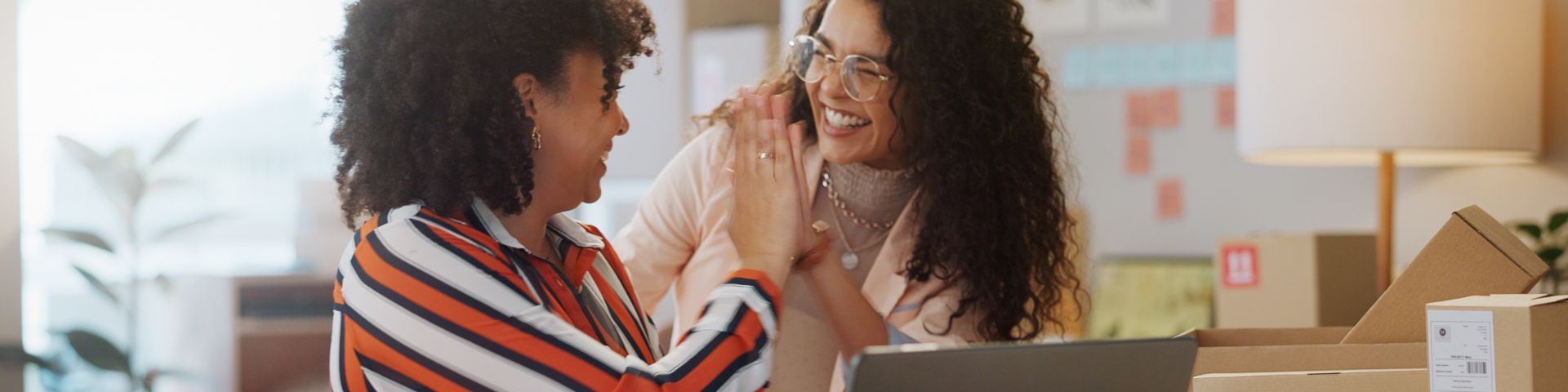 Duas mulheres sorrindo e comemorando juntas, uma delas usando óculos e ambas com cabelos cacheados, em um ambiente de escritório.