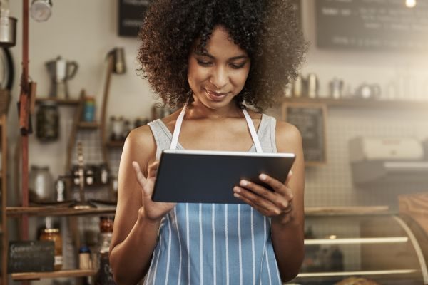 Mulher com cabelo crespo usando um tablet em um café, mostrando interesse em seu conteúdo. A cena reflete a modernidade e a tecnologia no ambiente de trabalho.