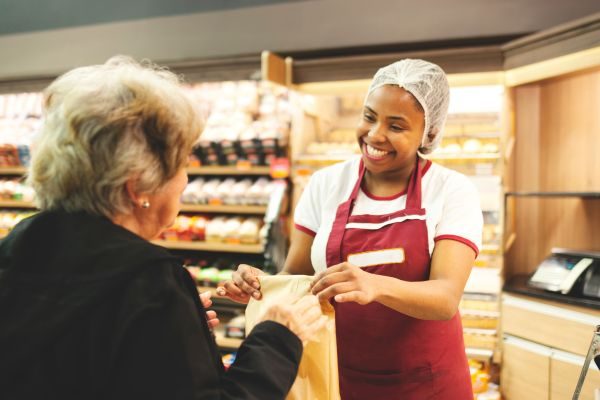 Funcionária sorridente entregando um pacote para uma cliente em uma padaria, mostrando atendimento ao cliente de qualidade e caloroso.