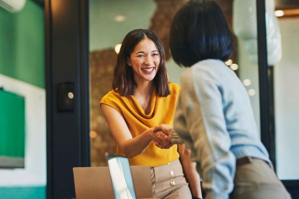 Duas mulheres sorrindo e se cumprimentando em um ambiente de trabalho moderno, simbolizando uma interação profissional positiva.