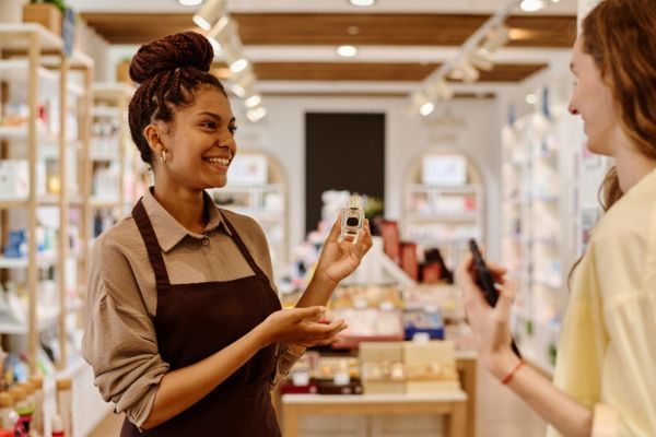 Uma vendedora sorridente demonstra um produto para uma cliente em uma loja bem iluminada, cercada por prateleiras de produtos de beleza.