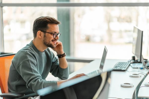 Homem em uma mesa de trabalho, falando ao celular enquanto navega no computador, em um ambiente de escritório moderno