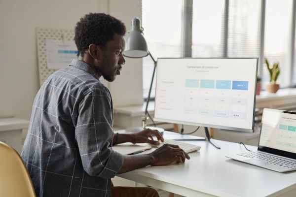 Homem trabalhando em um escritório moderno, analisando gráficos e tabelas no computador, representando produtividade e eficiência.