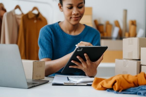 Mulher organizando invetário pelo tablet em um ambiente de trabalho com caixas e um laptop, simbolizando eficiência em gestão de estoques.