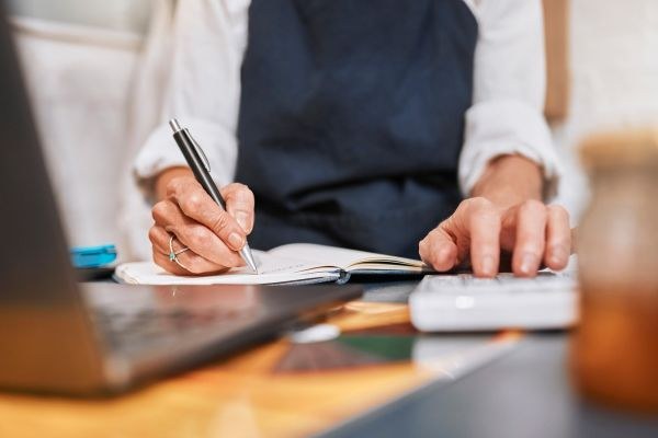 Pessoa escrevendo em um caderno enquanto utiliza um laptop e uma calculadora sobre a mesa, representando produtividade e organização.