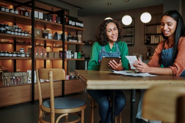 Duas empreendedoras com avental em uma loja, mexendo no tablet e conversando, simbolizando colaboração e gestão no ambiente de negócios