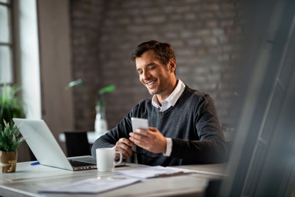 Homem sorridente sentado em uma mesa de trabalho, usando um smartphone e um laptop, cercado por anotações e uma xícara de café.