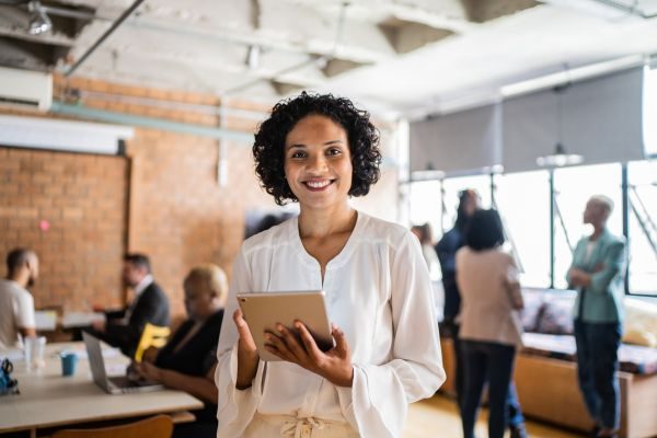 Mulher sorridente em ambientes de trabalho contemporâneo segurando um tablet, com colegas ao fundo, ideal para temas de produtividade e tecnologia.