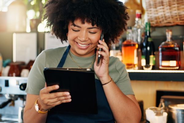 Mulher sorridente usando um tablet enquanto fala ao telefone em um ambiente de bar com bebidas ao fundo.