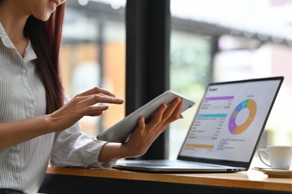 Mulher com foco nas mãos enquanto interage com um tablet e observa gráficos e dados em um notebook, em um ambiente de escritório