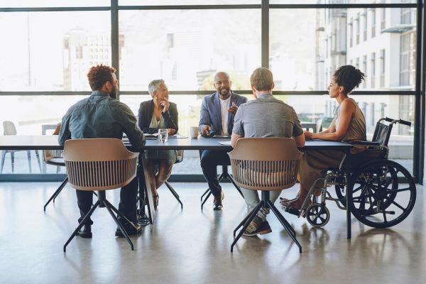 Grupo diverso em reunião ao redor de uma mesa em escritório iluminado, incluindo uma pessoa em cadeira de rodas, destacando inclusão e colaboração