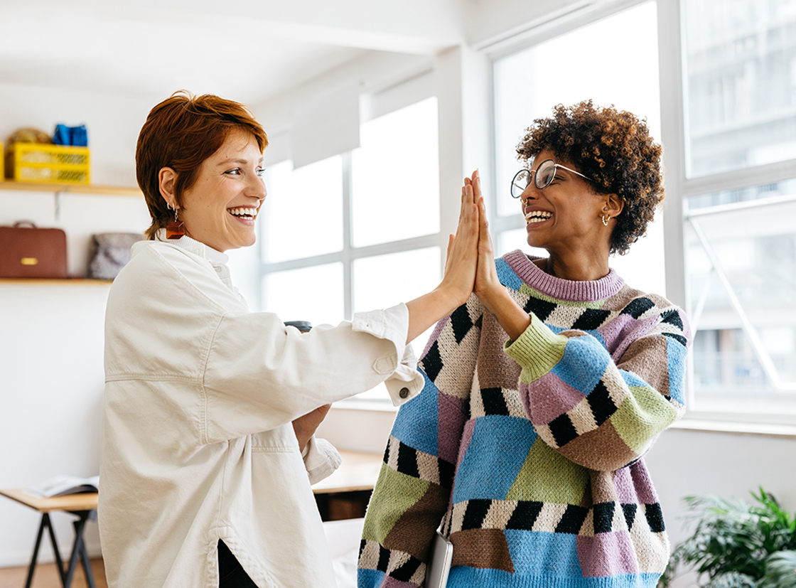 Duas mulheres diversas sorrindo e fazendo um high-five em um ambiente de escritório iluminado, transmitindo cooperação e positividade