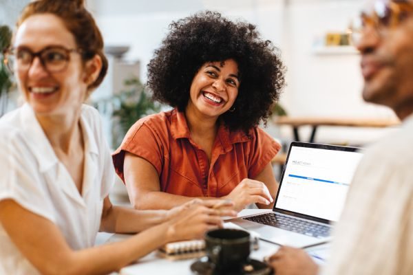 Mulher negra sorridente, com cabelo afro, participa de reunião ao lado de colegas, transmitindo confiança e liderança em ambiente colaborativo de trabalho.