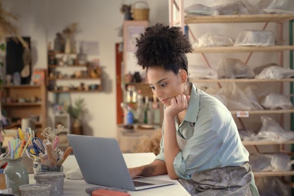 Mulher pensativa trabalhando em um computador em um ambiente criativo e artístico, mostrando sua dedicação e foco em projetos de cerâmica.