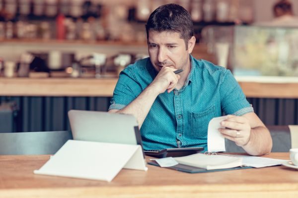 Homem sentado, segurando um papel em uma mão e olhando para um tablet, em um ambiente de trabalho.