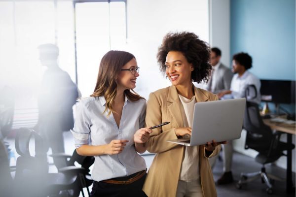 Duas mulheres jovens em um ambiente de escritório moderno, sorrindo e discutindo sobre um laptop. Elas representam o trabalho em equipe e a colaboração profissional.