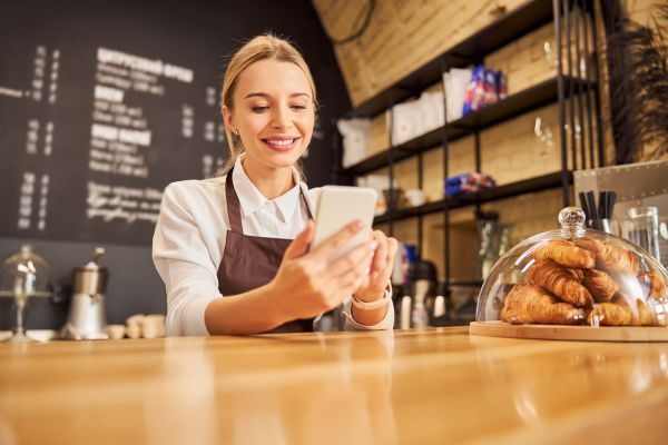 Imagem de uma funcionária de cafeteria sorrindo enquanto segura um celular, com croissants visíveis em uma vitrine ao fundo.