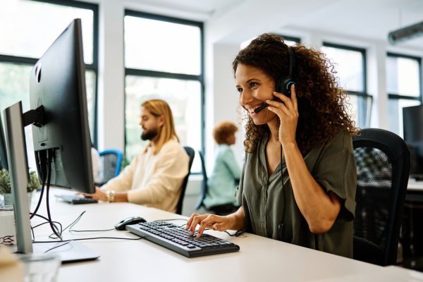 Mulher sorridente de telemarketing falando no telefone, usando fones de ouvido, com um computador à sua frente.