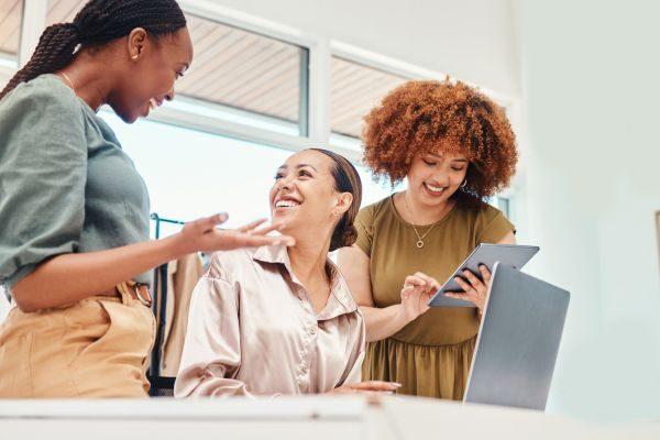 Três mulheres em ambiente colaborativo, discutindo ideias e usando tablet e laptop, destacando a dinâmica do trabalho em equipe.