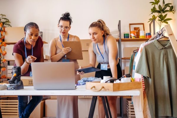 Três mulheres analisam informações em um laptop em um ambiente de trabalho moderno, cercadas por itens de vestuário e ferramentas de organização.