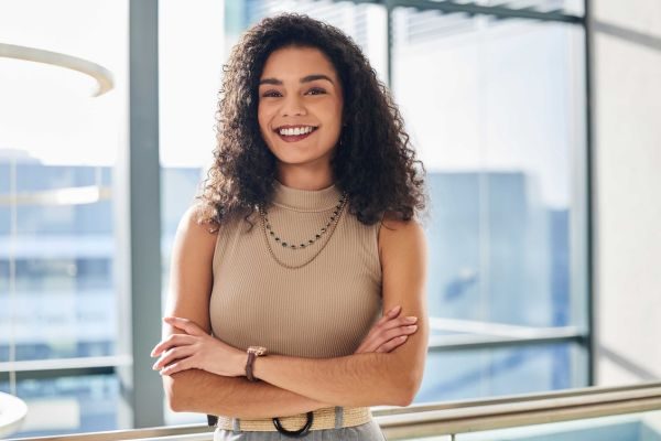 Mulher com cabelo cacheado, vestindo blusa bege, sorrindo e com os braços cruzados em ambiente moderno. Essa imagem retrata confiança e profissionalismo.