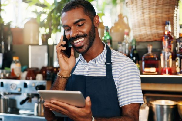 Homem sorridente em um bar, falando ao telefone e usando um tablet, transmitindo um ambiente descontraído e acolhedor.