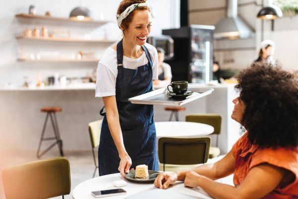 Imagem de uma garçonete servindo café e bolo para uma cliente em um café, com ambas sorrindo durante a interação.