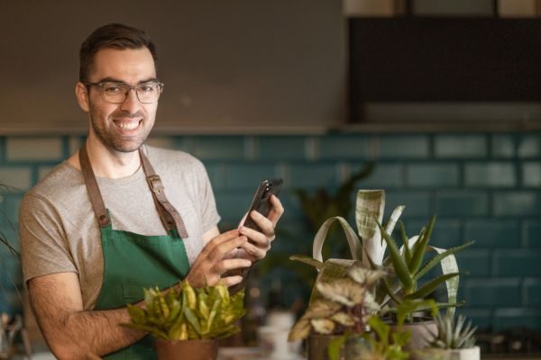 Homem sorridente trabalhando em uma loja de plantas, usando um avental verde e segurando um celular, rodeado por diversas plantas em um ambiente moderno.