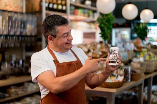 Homem sorridente em loja, mostrando celular com imagens de produtos. O ambiente é agradável, repleto de itens frescos e organizados.