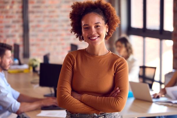 Mulher sorrindo em ambiente de escritório moderno, destacando-se com a sua confiança e simpatia, ideal para uso em contextos profissionais e de trabalho em equipe.