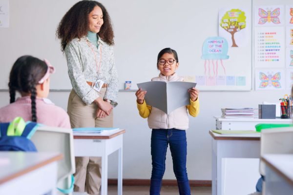 Criança apresentando trabalho escolar para a turma, sob a supervisão de uma professora em sala de aula. Ambiente educacional positivo e motivador.