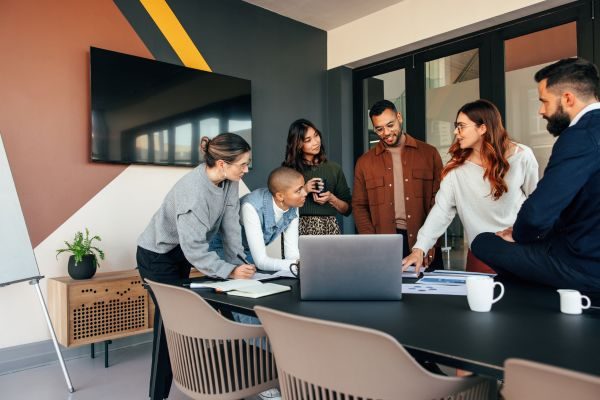 Equipe diversa em reunião colaborativa ao redor de uma mesa em escritório moderno, com laptop e anotações, demonstrando trabalho em equipe