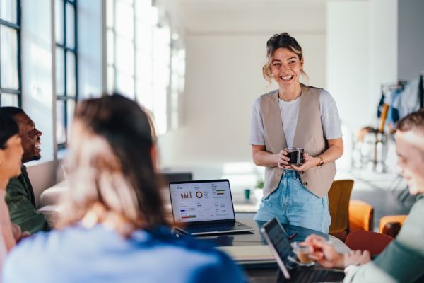 Quatro pessoas em uma reunião colaborativa em um ambiente moderno, com foco em uma mulher sorridente apresentando ideias. Eles estão ao redor de uma mesa com laptops e gráficos.