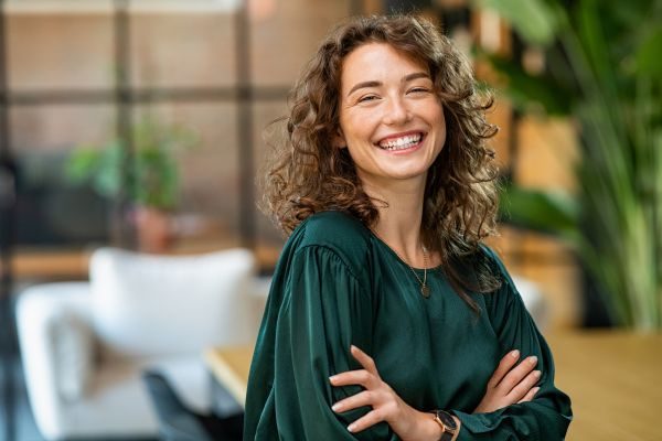 Mulher sorridente com cabelo cacheado e blusa verde em ambiente moderno, expressando alegria e confiança. Ideal para ilustrar felicidade e bem-estar.