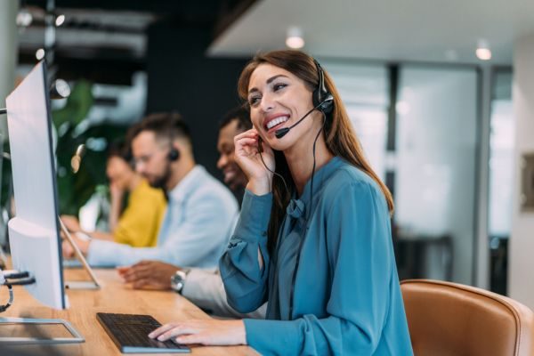 Atendente feliz em call center com fones de ouvido, sorri enquanto auxilia clientes em um escritório moderno com outros colegas ao fundo.