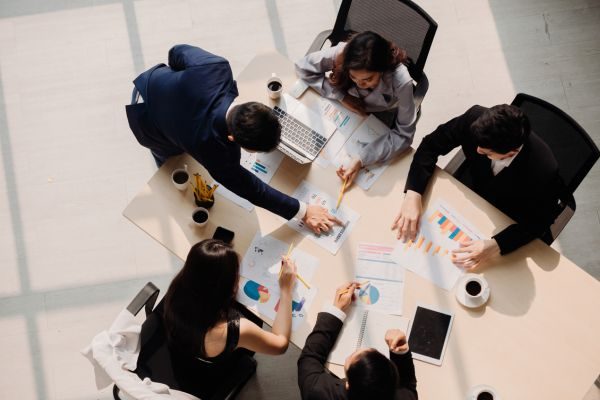 Grupo de pessoas em reunião ao redor de uma mesa, analisando gráficos e discutindo estratégias, com laptops, relatórios e xícaras de café visíveis.