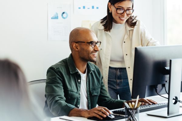 Homem sentado em um ambiente de trabalho, olhando para o computador, com uma mulher em pé ao lado dele, ambos focados na tela.