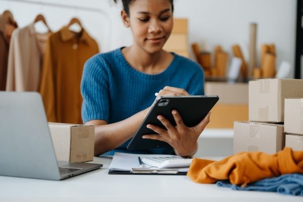 Empreendedora usando um tablet para organizar pedidos, cercada por caixas, roupas e um laptop em um ambiente de trabalho.