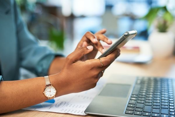 Uma mulher utilizando um celular enquanto trabalha em um laptop, mostrando a interatividade entre dispositivos na rotina profissional.