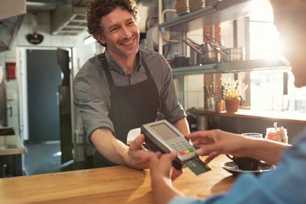 Atendente de restaurante sorridente entregando uma máquina de cartão para pagamento, em um balcão de ambiente iluminado e acolhedor.