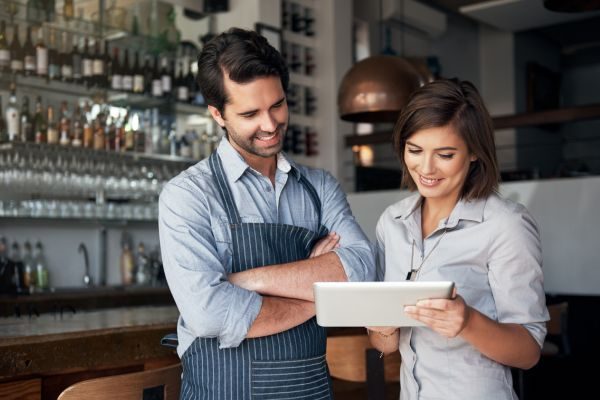 Homem de avental e mulher de roupa social analisando informações em um tablet juntos, em um ambiente de trabalho.