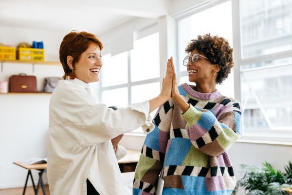 Duas mulheres diversas sorrindo e fazendo um high-five em um ambiente de escritório iluminado, transmitindo cooperação e positividade