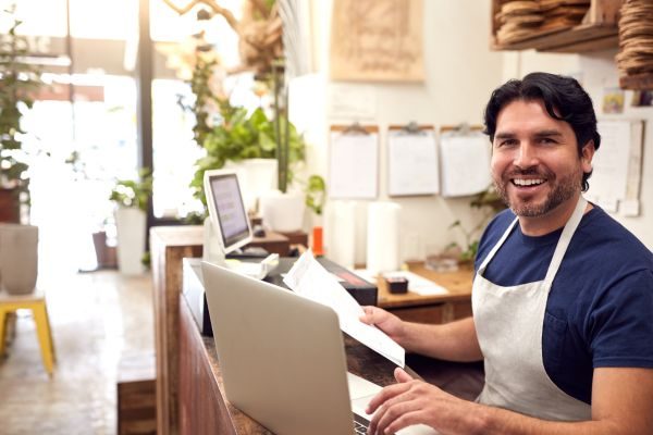 Empresário com avental sorrindo e mexendo no computador sozinho, representando satisfação profissional.