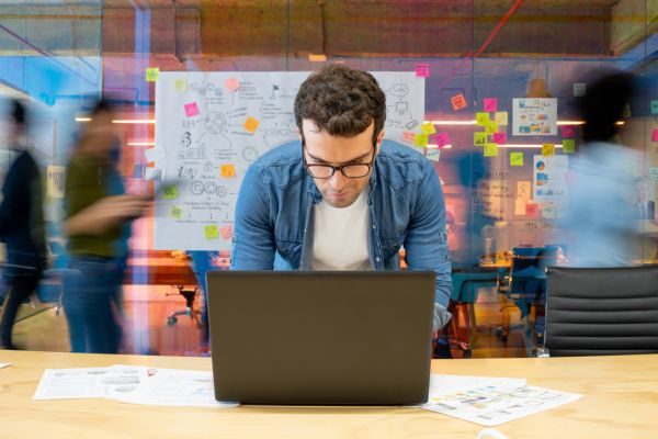 Homem concentrado em trabalho remoto em um laptop, rodeado por um ambiente moderno e criativo com equipes colaborando ao fundo.