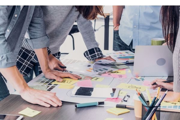 Pessoas reunidas em uma mesa de trabalho, rodeadas por post-its, papéis e materiais de escritório, colaborando em um ambiente dinâmico.