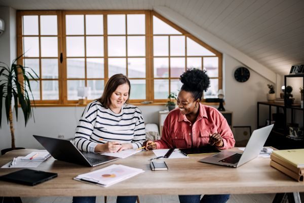 Dupla de mulheres trabalhando em um ambiente de escritório moderno, sorrindo e interagindo enquanto revisam documentos e usam laptops.