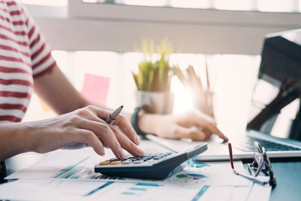 Mão segurando uma caneta e mexendo na calculadora, com óculos de canto e um computador ao fundo, simbolizando foco em finanças e organização