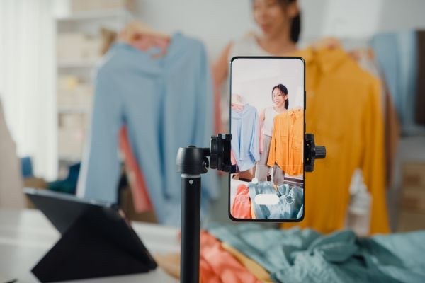 Uma modelo apresenta roupas coloridas em um vídeo gravado com um smartphone, enquanto se destaca um ambiente de moda moderno e organizado.