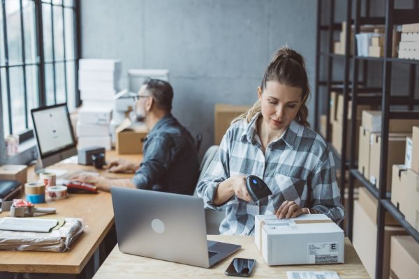 Pessoas trabalhando em um pequeno negócio: uma escaneia uma caixa enquanto a outra trabalha no computador, com itens de embalagem sobre a mesa.