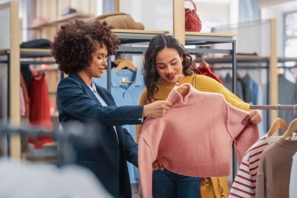 Duas mulheres sorrindo em uma loja de roupas, analisando um suéter rosa, destacando a experiência de compra e a moda feminina.